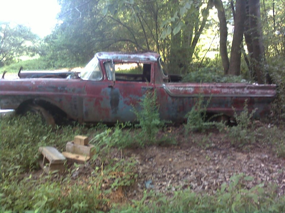 Derelict faded red car-based pickup with missing glass sitting in weeds at wood's edge