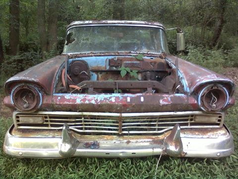 1973 Ford Ranchero - Head-on view of a derelict 1957 Ford sitting in tall grass in the woods, hood and engine gone, with empty headlight buckets, hanging wires,…
