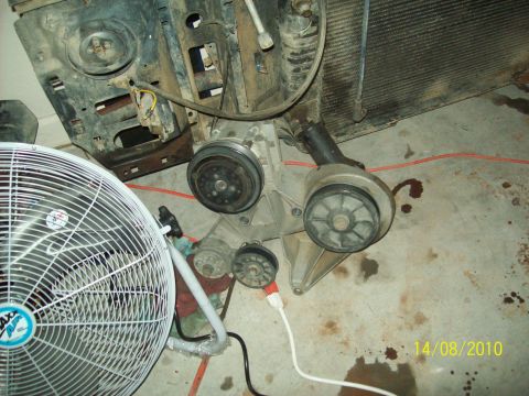 1985 Ford Bronco - Disassembled engine components, including a bracket with pulleys and a compressor clutch, are laid out on a concrete floor.