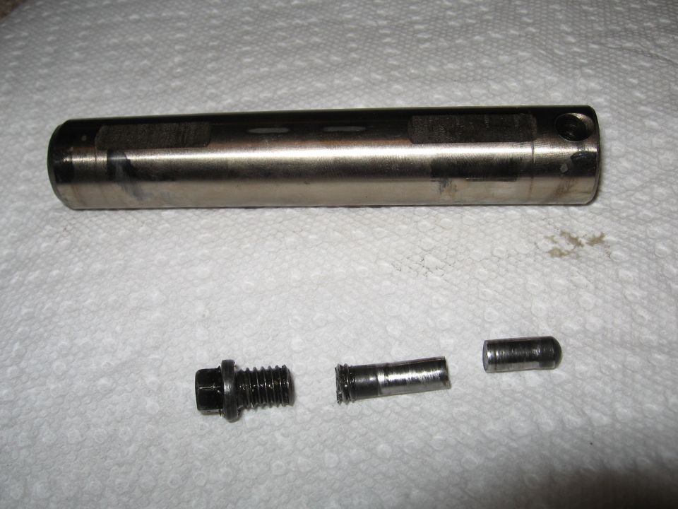 Metal automotive part, possibly a pin or shaft, with a bolt and two smaller cylindrical pieces laid out on paper towels