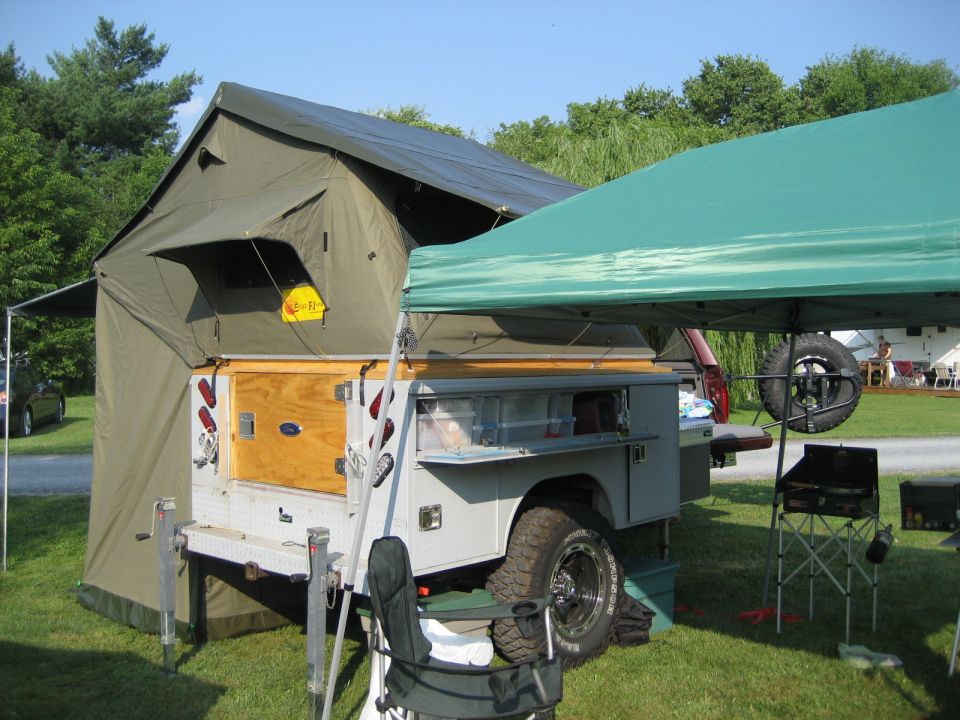 Off-road trailer with a large tent set up, a spare tire mounted on the back, and a green canopy providing shade