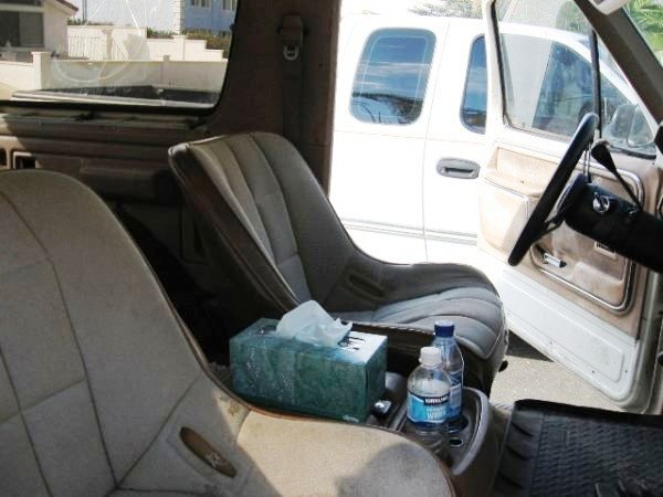 Interior of a vehicle showing bucket seats, a tissue box, and water bottles. A white truck is visible through the open door