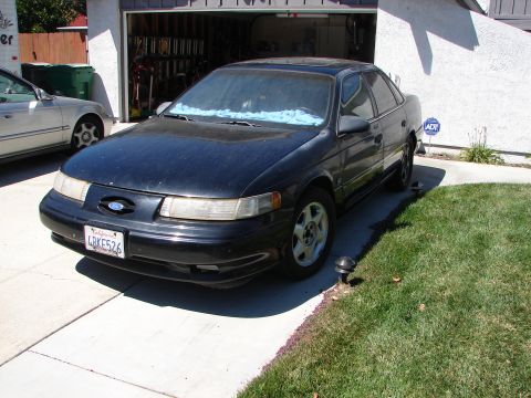 1992 Ford Taurus - A black 1992 Ford Taurus sedan is parked in a driveway next to another vehicle.