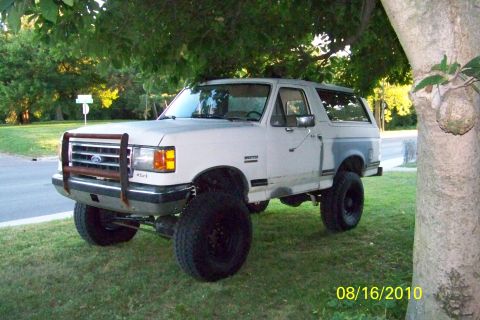 1989 Ford Bronco - Three-quarter front view of a lifted white Ford Bronco with a removable hardtop, parked on grass under a shade tree at dusk. It wears a rus…