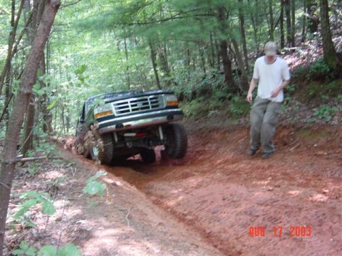 1995 Ford Bronco - Dark green Ford Bronco leaning hard off-camber with its driver-side front tire dropped into a washed-out ditch on a red-clay forest trail,…