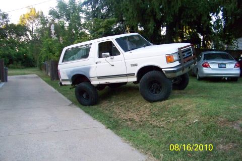 1989 Ford Bronco - A white 1989 Ford Bronco with oversized tires is parked on a grassy slope next to a driveway, with another car visible in the background.