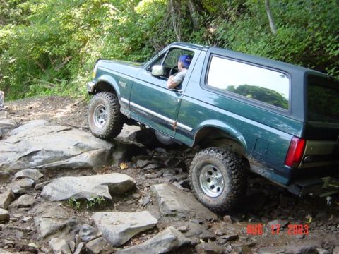 1995 Ford Bronco - Dark green full-size Ford Bronco with removable hardtop crawling nose-down over a rocky ledge on a wooded trail, seen from the driver's sid…