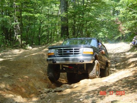 1995 Ford Bronco - Head-on front view of a dark green lifted Ford Bronco climbing out of a deep dirt rut on a wooded trail, front tires articulated over the u…
