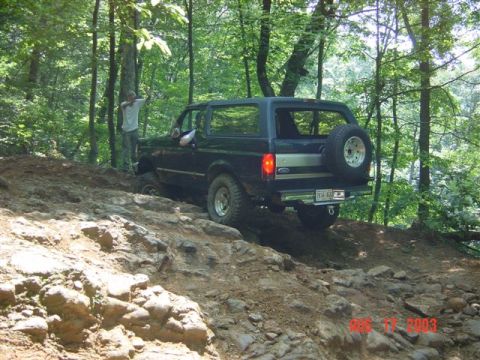 1995 Ford Bronco - Rear three-quarter view of a dark green full-size Ford Bronco with removable hardtop climbing a rocky, rutted trail through the woods, body…