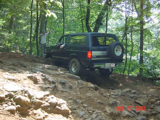 Dark green Ford Bronco climbing a rocky wooded trail, rear three-quarter view with spare tire