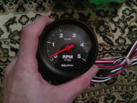 1990 Ford Bronco - A hand holds a black Auto Meter tachometer with a red needle, designed for diesel engines.