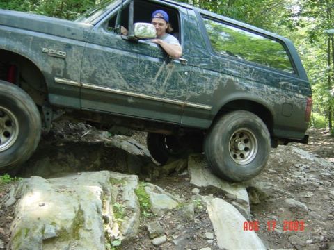 1995 Ford Bronco - Driver's-side view of a mud-streaked dark green full-size Ford Bronco flexing over a rock ledge on a wooded trail, with the front driver's…