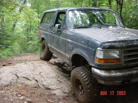 1995 Ford Bronco - Low passenger-side front three-quarter view of a mud-splattered dark green Ford Bronco descending a slab of exposed rock on a wooded trail.…