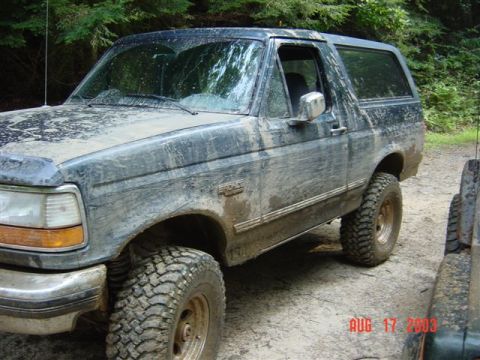 1995 Ford Bronco - Mud-splattered dark green Ford Bronco full-size SUV with removable hardtop, photographed from the driver's front three-quarter angle on a g…