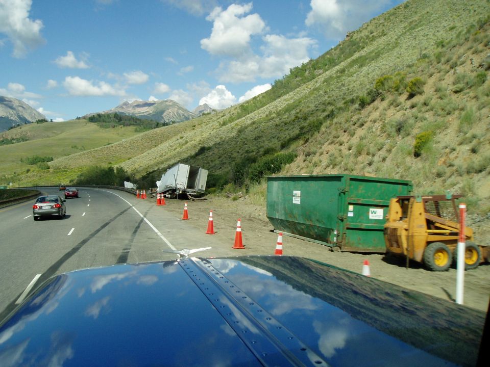 Roadside accident scene with overturned trailer, green dumpster, and yellow skid steer loader. Cars drive past on the highway