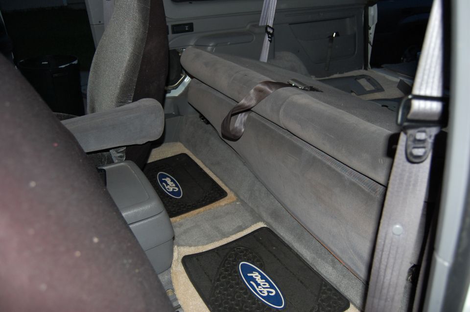 Interior of a vehicle showing grey seats, floor mats with Ford logos, and a seatbelt