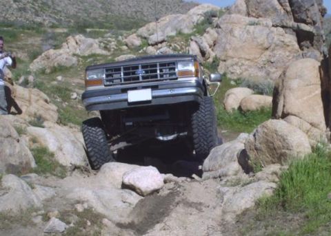 1990 Ford Bronco II - A 1990 Ford Bronco II is shown off-roading on a rocky trail with a person standing in the background.