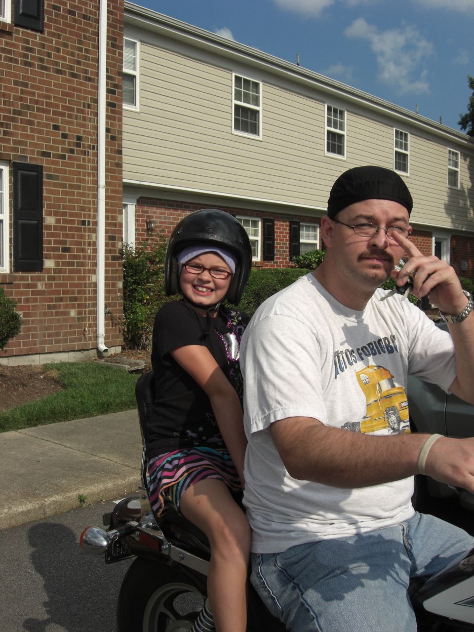 Man and young girl wearing helmets on a motorcycle, parked in front of townhouses