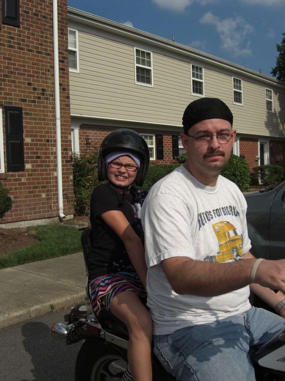 Man and young girl wearing helmets ride on a motorcycle in front of townhouses