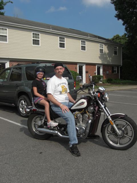 1986 Honda Shadow Aero - A man and child sit on a maroon motorcycle parked in front of an apartment building and an SUV.