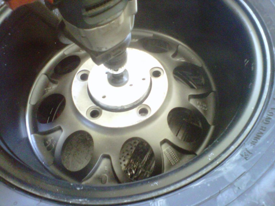 Drill bit working on a silver alloy wheel with black tire. Supermotors.net logo visible