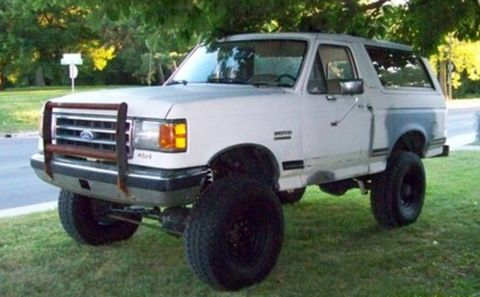 1989 Ford Bronco - Three-quarter front view of a lifted white late-1980s Ford Bronco parked on grass under trees, riding on oversized mud-terrain tires and bl…