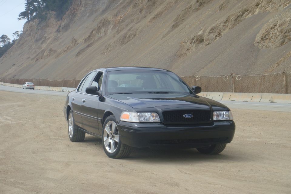 Black Ford Crown Victoria sedan, front three-quarter view, parked on dirt beside a highway hillside