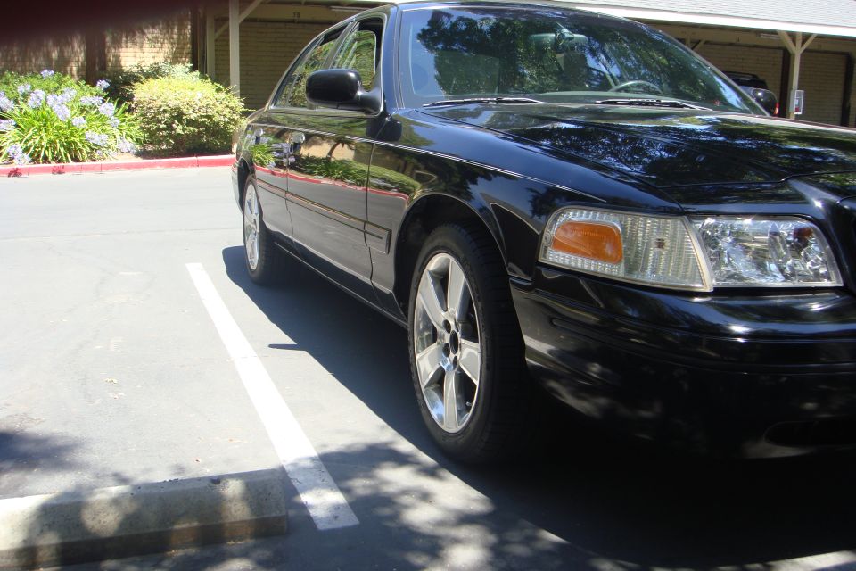 Black Crown Victoria sedan with five-spoke alloy wheels, front passenger-side view in a shaded parking lot