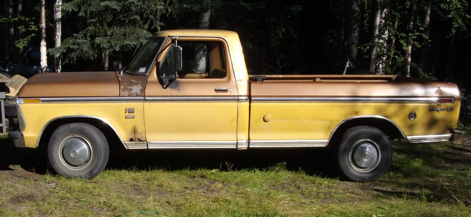 Side view of a 1976 Ford F-150 pickup truck, two-tone yellow and brown paint, parked in a grassy area with trees behind it