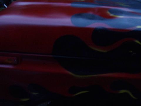 1978 Ford Bronco - Dim close-up of a red front fender and hood painted with black flames outlined in yellow, focused on the seam where the hood meets the fend…