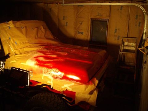 1978 Ford Bronco - A vehicle hood, mostly covered by a yellow tarp, is being painted red with visible paint splatters.