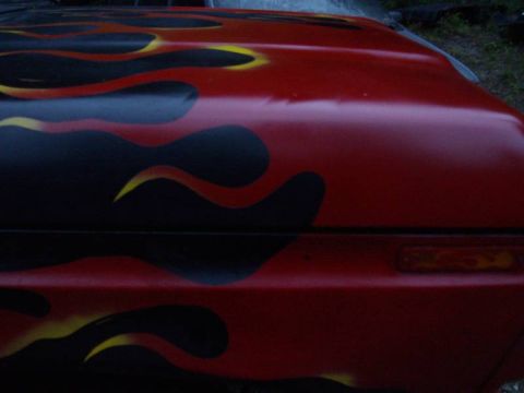 1978 Ford Bronco - Vehicle hood painted with red, black, and yellow flames. Part of a headlight is visible on the right.