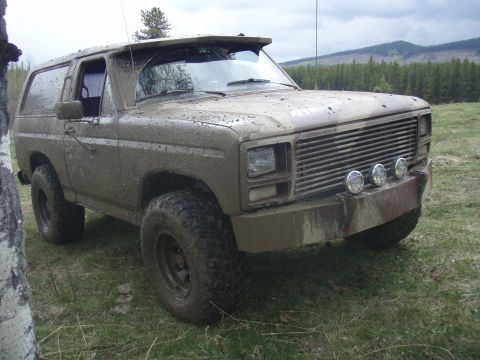 1985 Ford Bronco - This heavily muddied SUV, likely from the early 1980s, is shown in a rural outdoor setting.