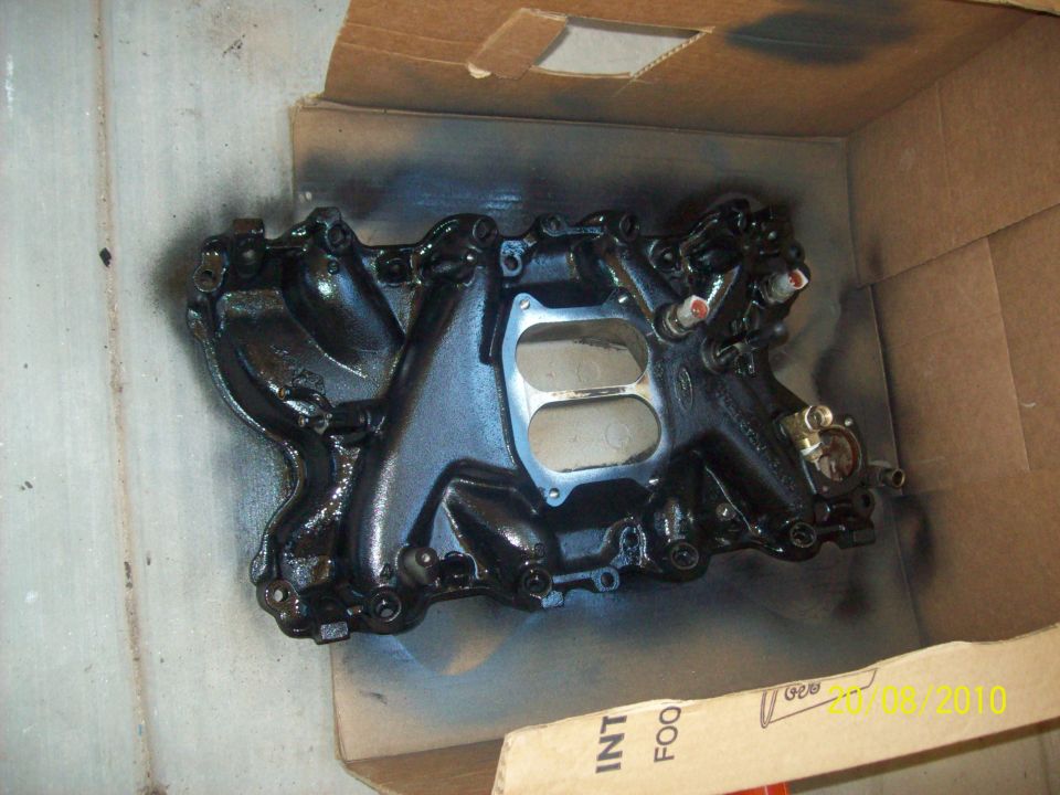 Black intake manifold with dual carburetor openings, inside a cardboard box