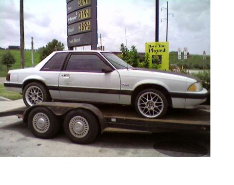 1979 Ford F-150 - A white coupe with a black hood is transported on a trailer near a gas station.