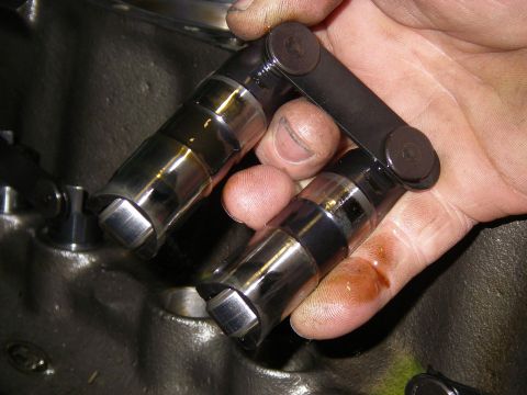 1990 Ford Bronco - A greasy hand holds a pair of new roller lifters joined by their link bar, the polished barrels still wet with assembly oil, above a bare e…