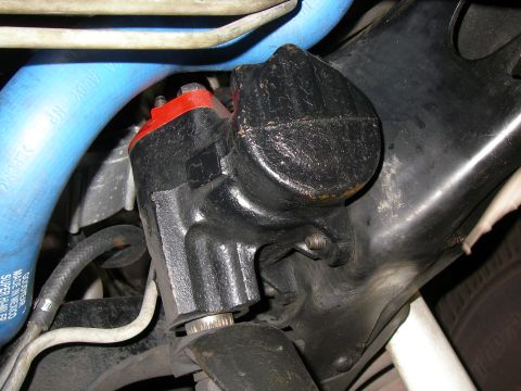 1990 Ford Bronco - Close-up of a power steering gearbox painted black with a red top cap, bolted to a black frame rail inside a wheel well area. Blue rubber c…