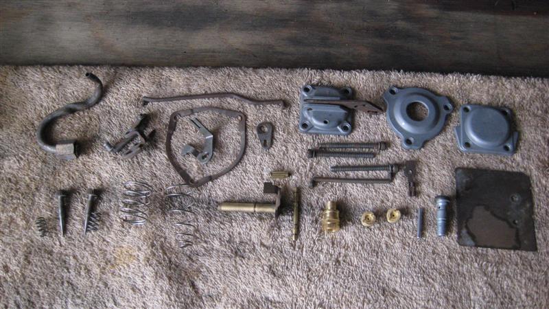 Disassembled carburetor parts laid out on a textured surface. Includes springs, metal levers, brass fittings, and housing components