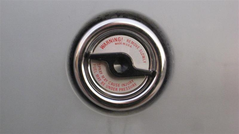 Close-up of a chrome fuel filler cap with a warning label about fuel spray