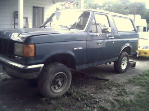 1989 Ford Bronco - A dark blue Ford Bronco is parked on a gravel driveway with a yellow car in the background.