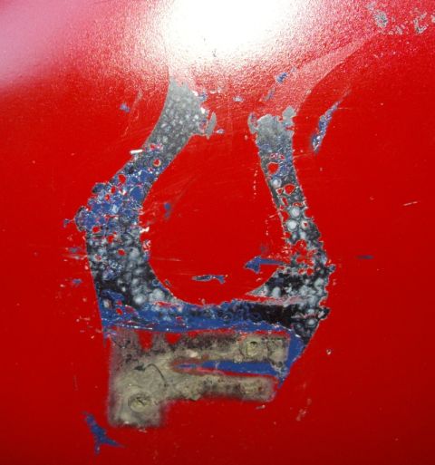 1978 Ford Bronco - Extreme close-up of a glossy red painted panel where a horseshoe-shaped emblem or bracket has been removed, leaving blistered blue and gray…