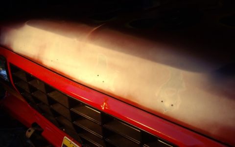 1978 Ford Bronco - Low-angle close-up of a red front end above an egg-crate grille, with harsh light raking across the upper panel to reveal skim-coat filler,…