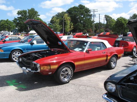 1968 Shelby GT500 - A red 1969 convertible with a white top and yellow side stripe, identified by a GT350 badge, is displayed with its hood open at a car show.
