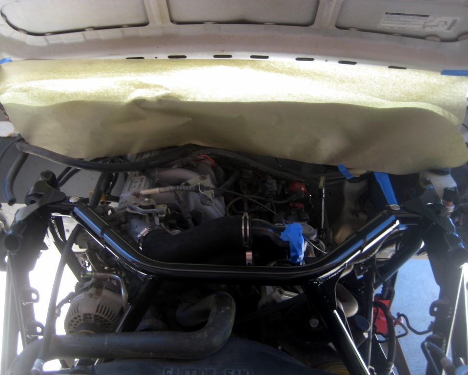 Engine bay with a black aftermarket brace installed over the engine. Some parts are covered with protective paper