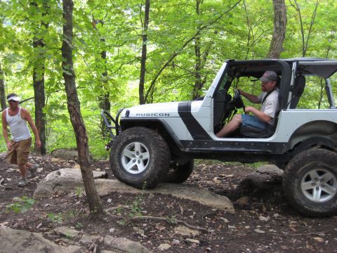 2005 Jeep Wrangler - A man drives a white Jeep Wrangler Rubicon through a rocky, wooded area, with another person watching.