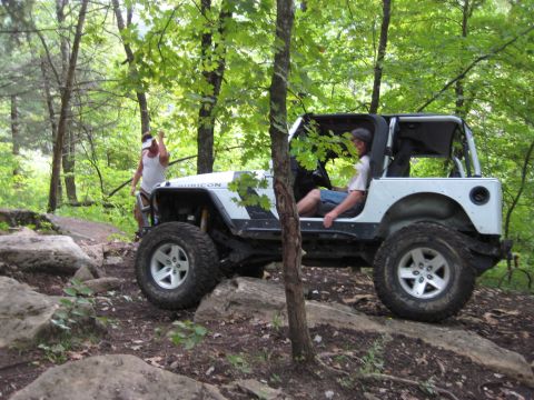 2005 Jeep Wrangler - Side view of a white Jeep Wrangler Rubicon with doors and top removed climbing a rocky ledge on a shaded forest trail, front tire up on a r…