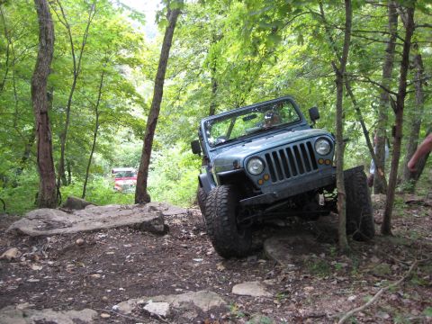 2005 Jeep Wrangler - A gray-green topless Jeep Wrangler TJ with the windshield up and doors off works down a rocky descent on a green wooded trail, front tires…