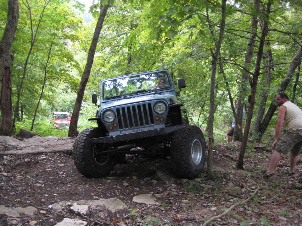 Blue-gray topless Jeep Wrangler flexing over rocks on a wooded off-road trail