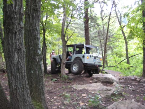 2005 Jeep Wrangler - Rear three-quarter shot of a light silver/white Jeep Wrangler with doors and top off, bare roll bar and tailgate-mounted spare, working a r…