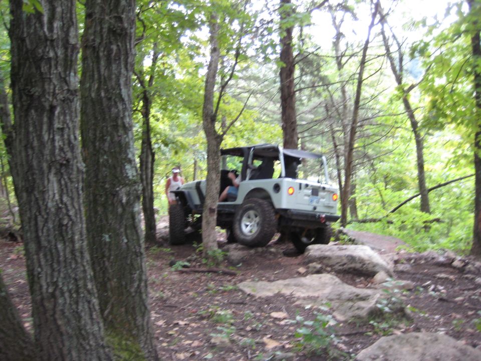 Silver doorless Jeep Wrangler on rocky forest trail with a spotter standing alongside
